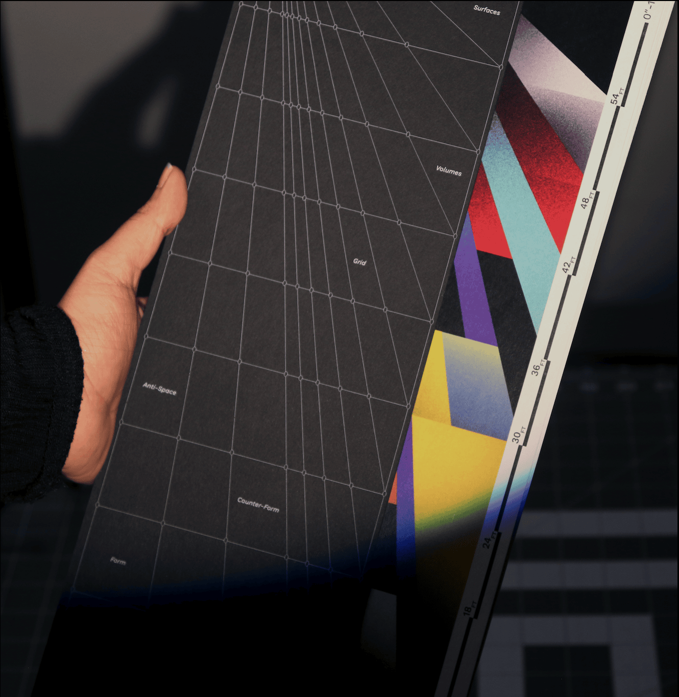 A hand holding the Thresholds book open on a perspective grid page, colour plates fanned behind it.
